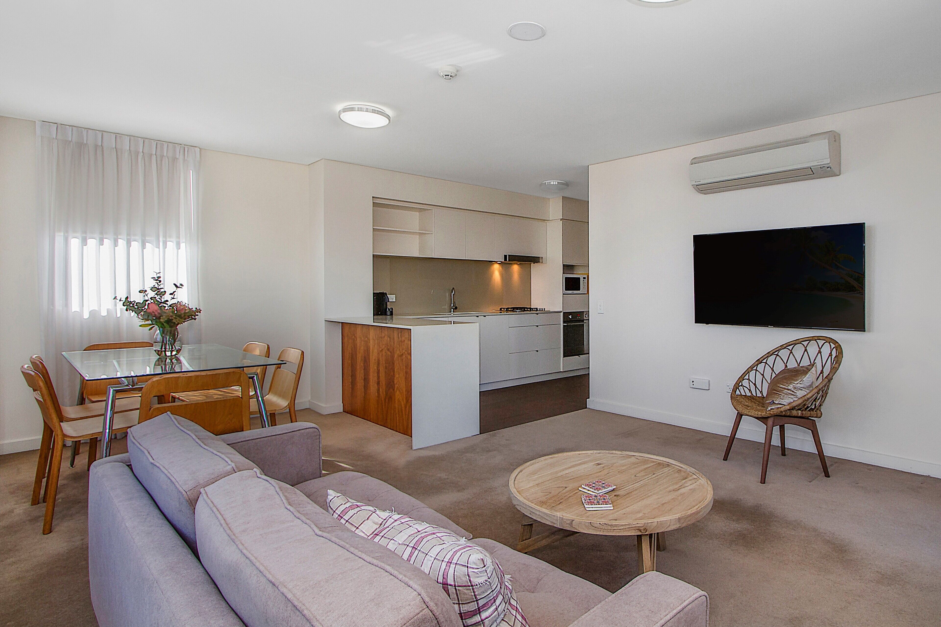 A modern living room with a sofa, coffee table, dining table, and chairs. A kitchen with white cabinets and a microwave is visible in the background.