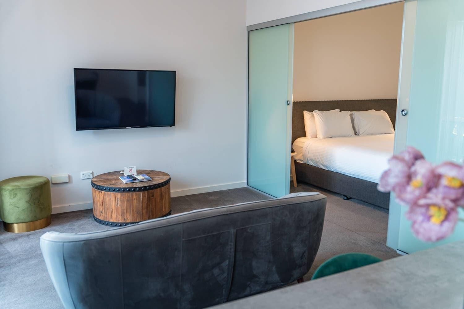 Een moderne hotelkamer met een flatscreen televisie, een rond houten bijzettafeltje, een grijze bank, een bed met witte lakens en een inloopkast met schuifdeuren.