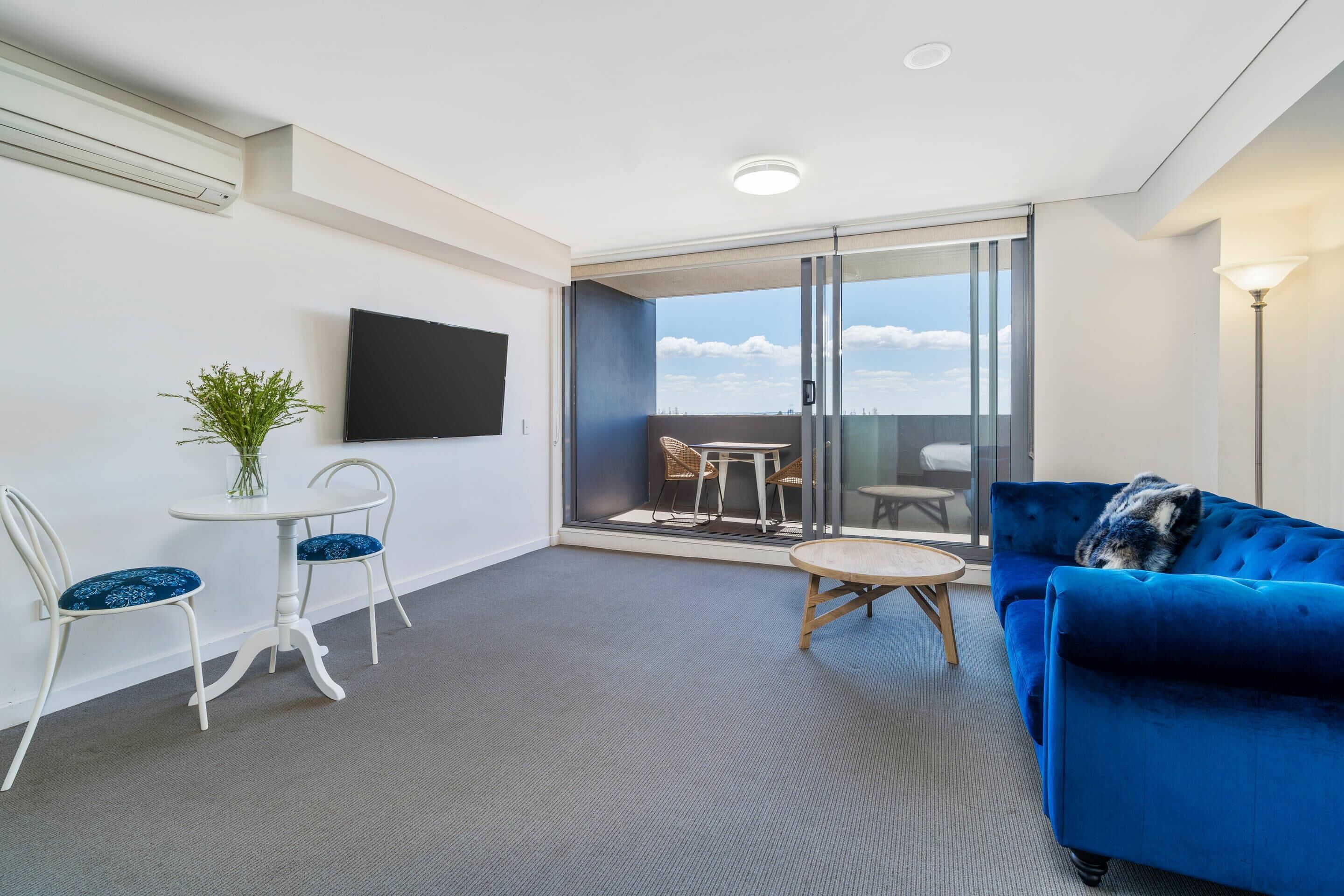 A modern living room with a blue sofa, two white chairs, a small round table, and a TV mounted on the wall.