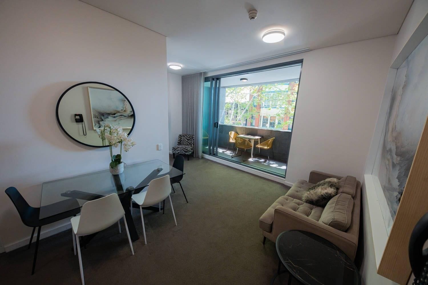 A modern living room with a glass dining table, a sofa, and a large window with a view of greenery.