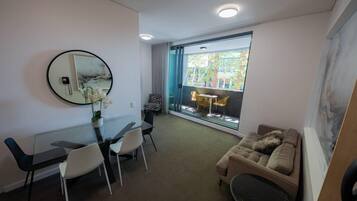 A modern living room with a glass dining table, a sofa, and a large window with a view of greenery.