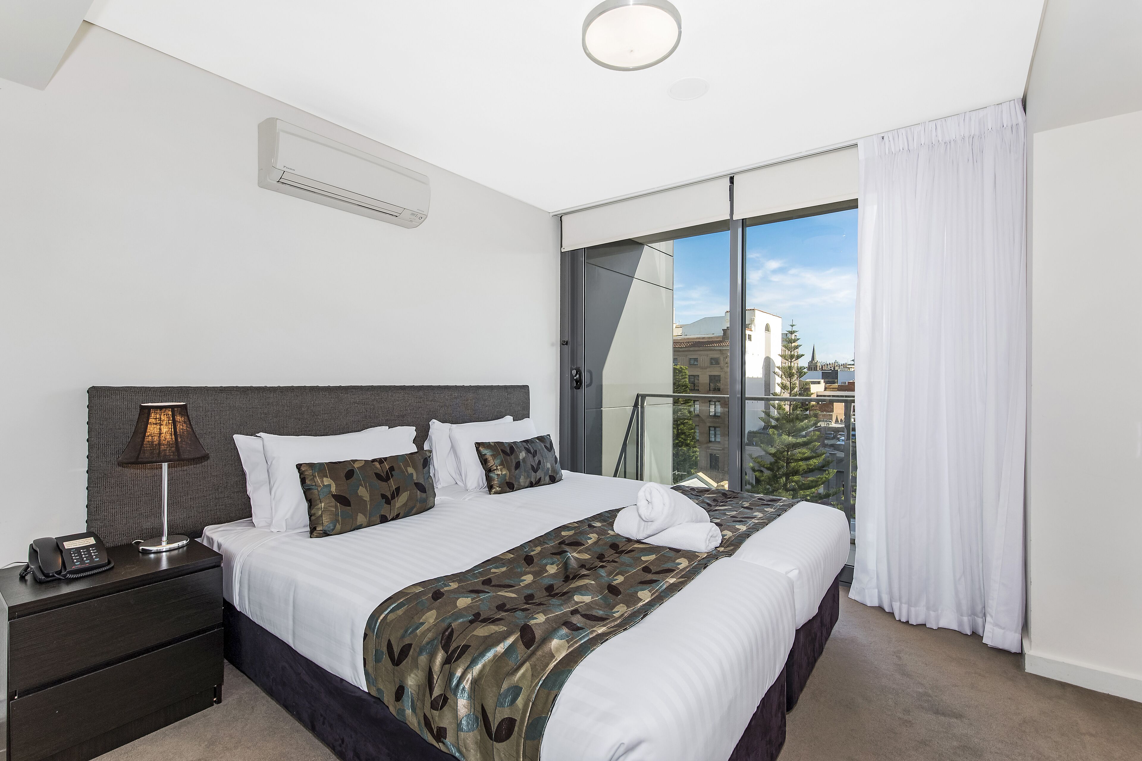 A modern hotel room with a large bed, a bedside table with a lamp, and a view of the city through a sliding glass door.