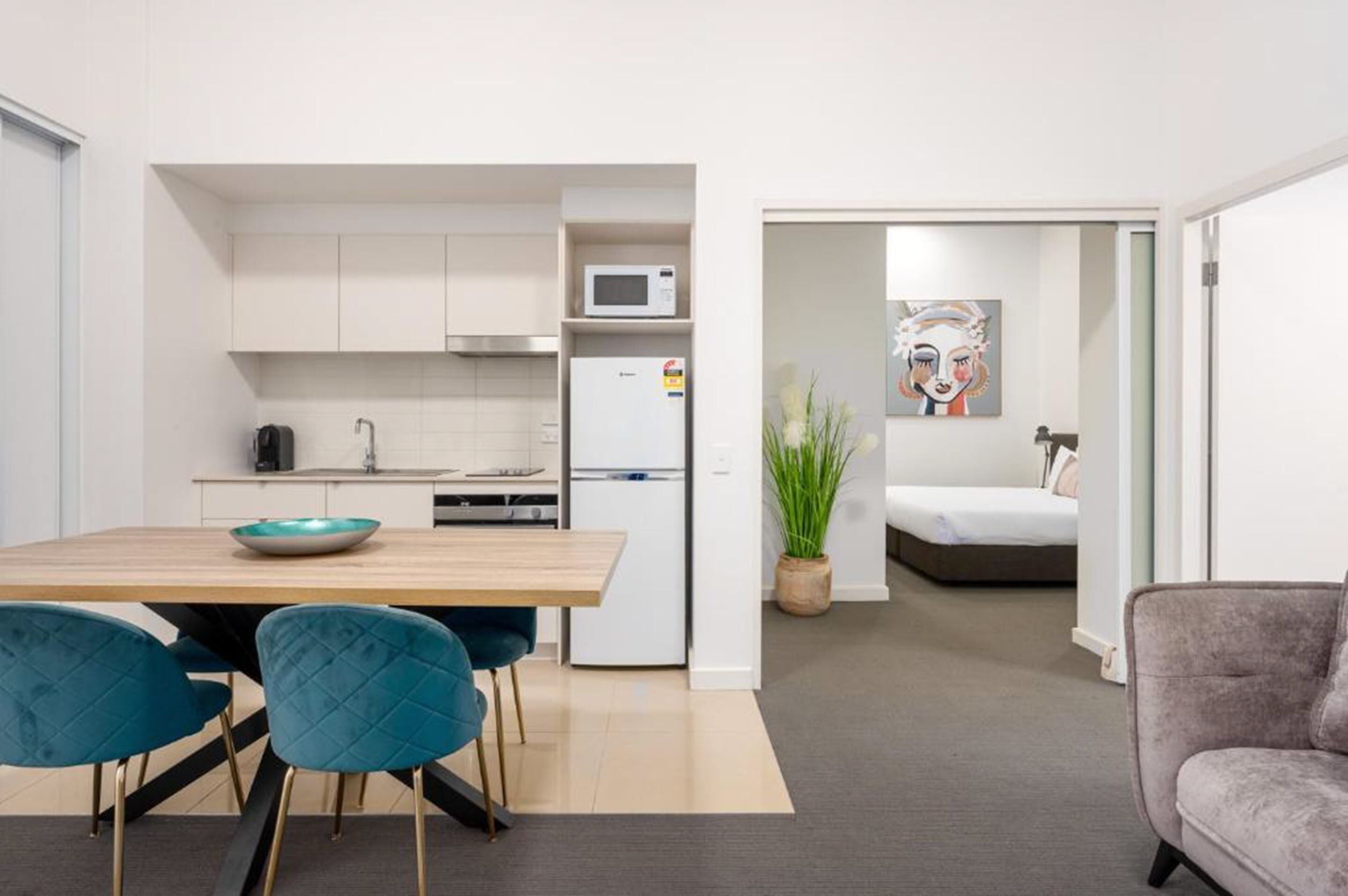A modern kitchen with white cabinets, a microwave, and a refrigerator. There is a dining table with chairs and a potted plant. A gray sofa is visible in the foreground.