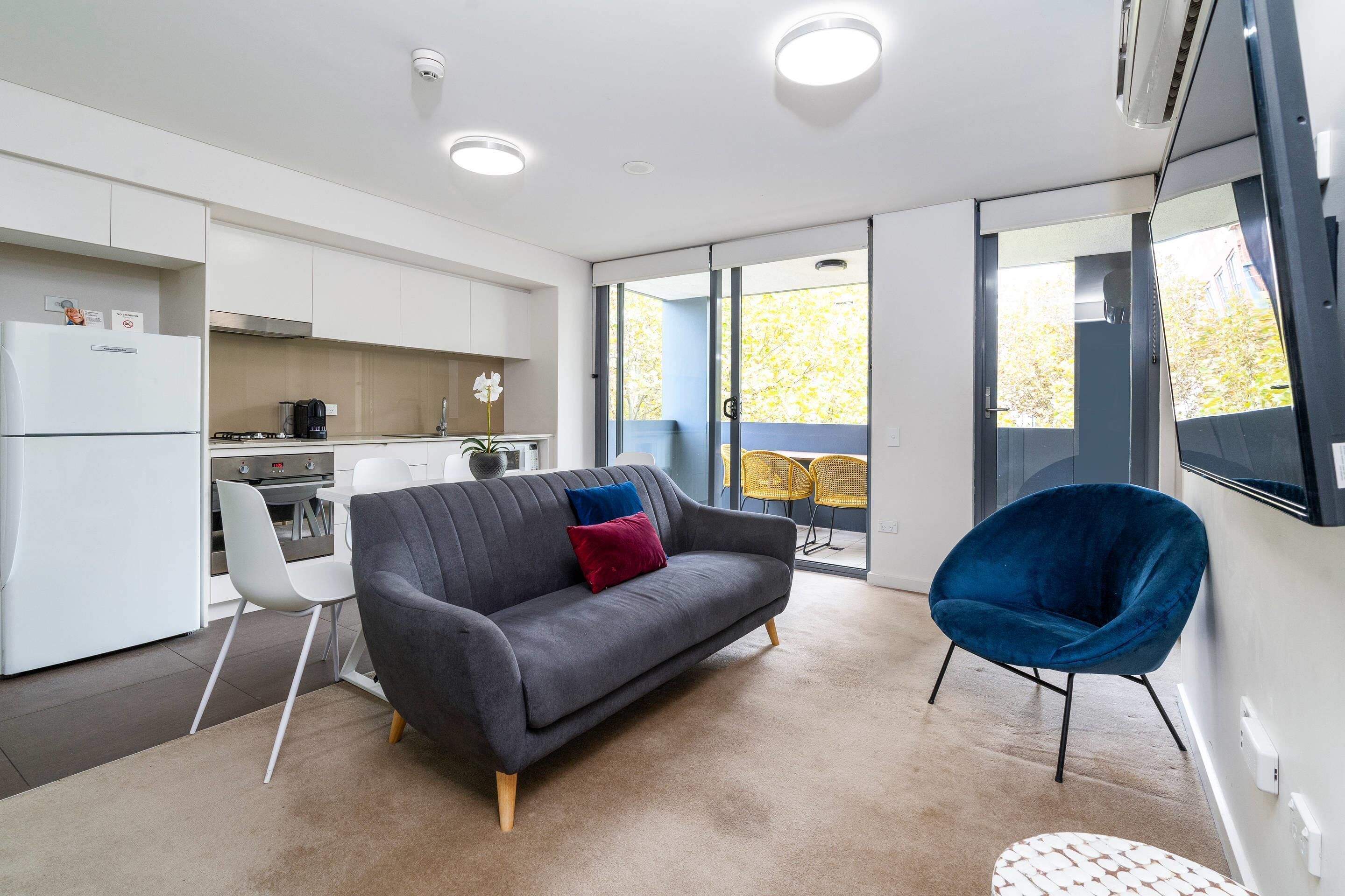 A modern kitchen with white cabinets, a grey sofa with colorful cushions, a blue chair, and a large window offering a view of trees.