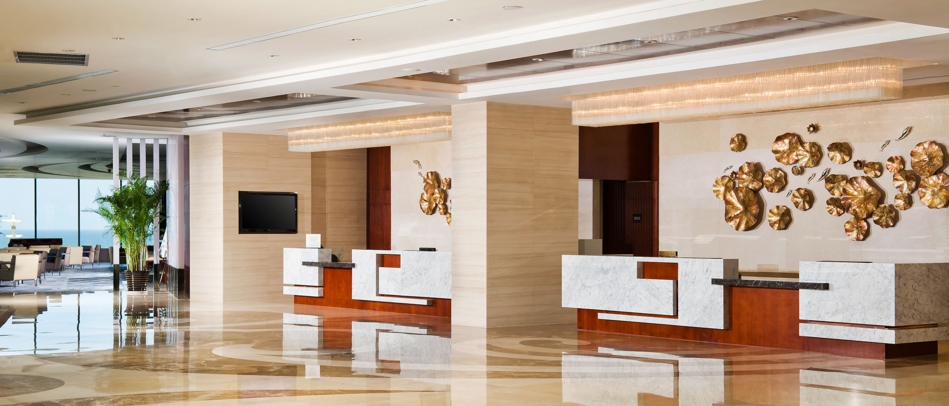 A modern hotel lobby with marble reception desks, a large gold decorative wall, and a view of the sea through large windows.