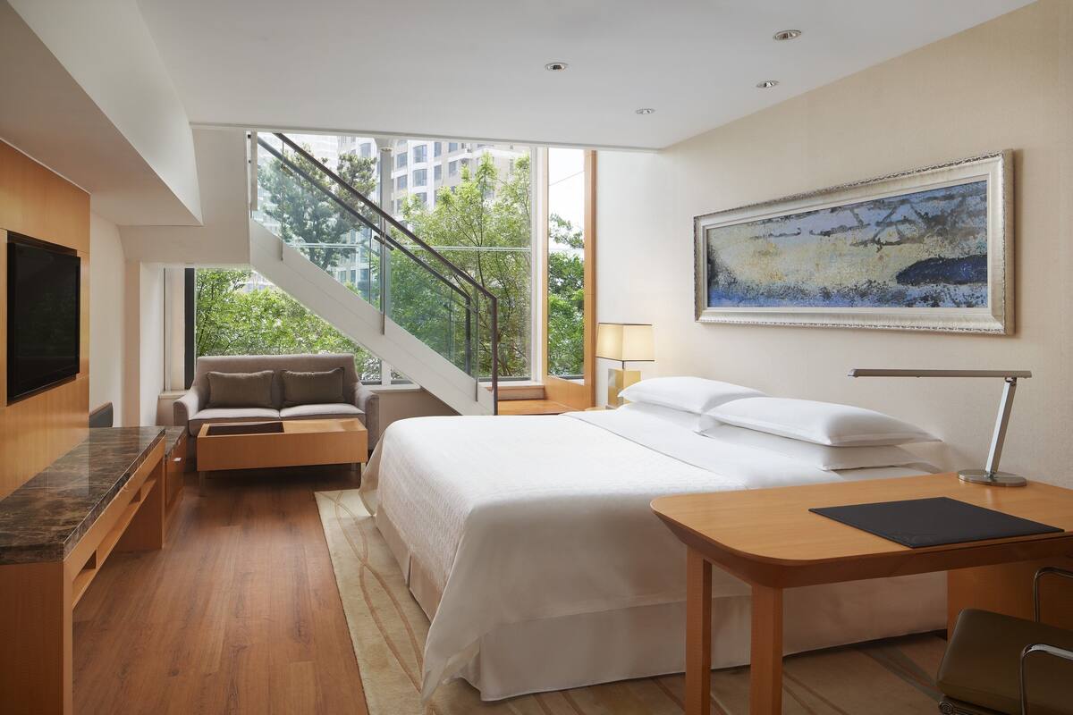 A modern hotel room with a large bed, a desk, a sofa, and a staircase leading to an outdoor area.
