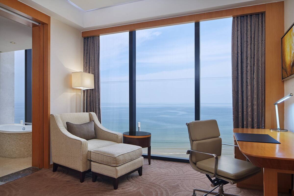 A hotel room with a large window, a desk, a chair, a sofa, and a small side table.