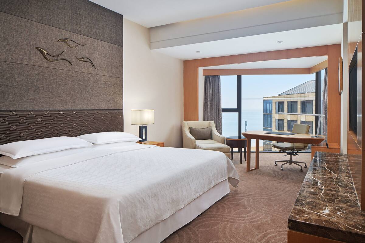 A hotel room with a large bed, a desk with a chair, a lamp, and a view of the city.
