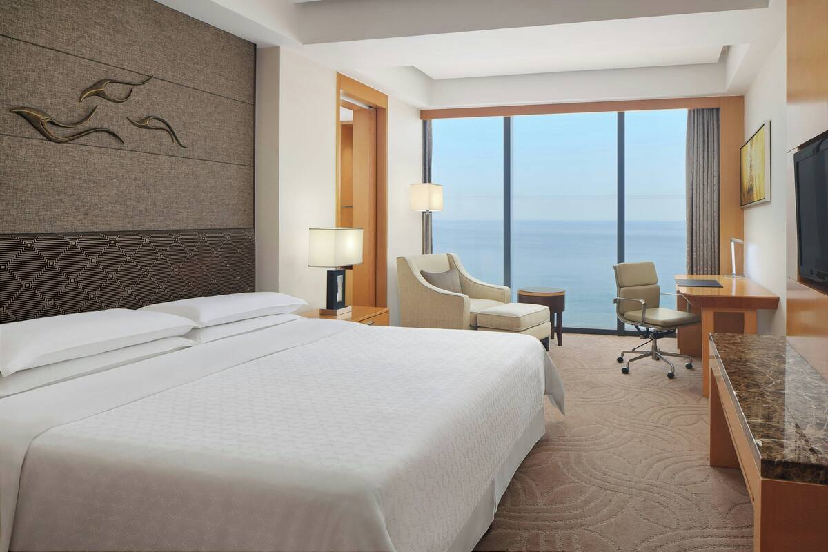 A hotel room with a large bed, two armchairs, a desk, a TV, and a view of the ocean.