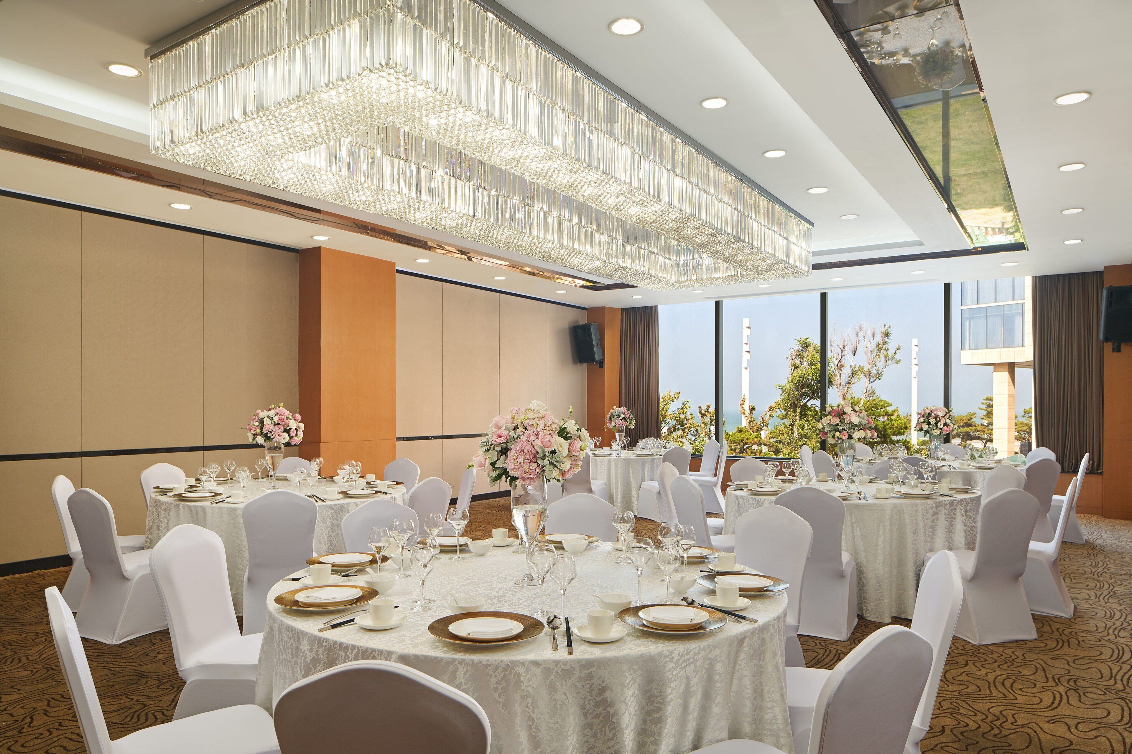 A banquet hall with round tables set for dining, white chairs, and a large chandelier.