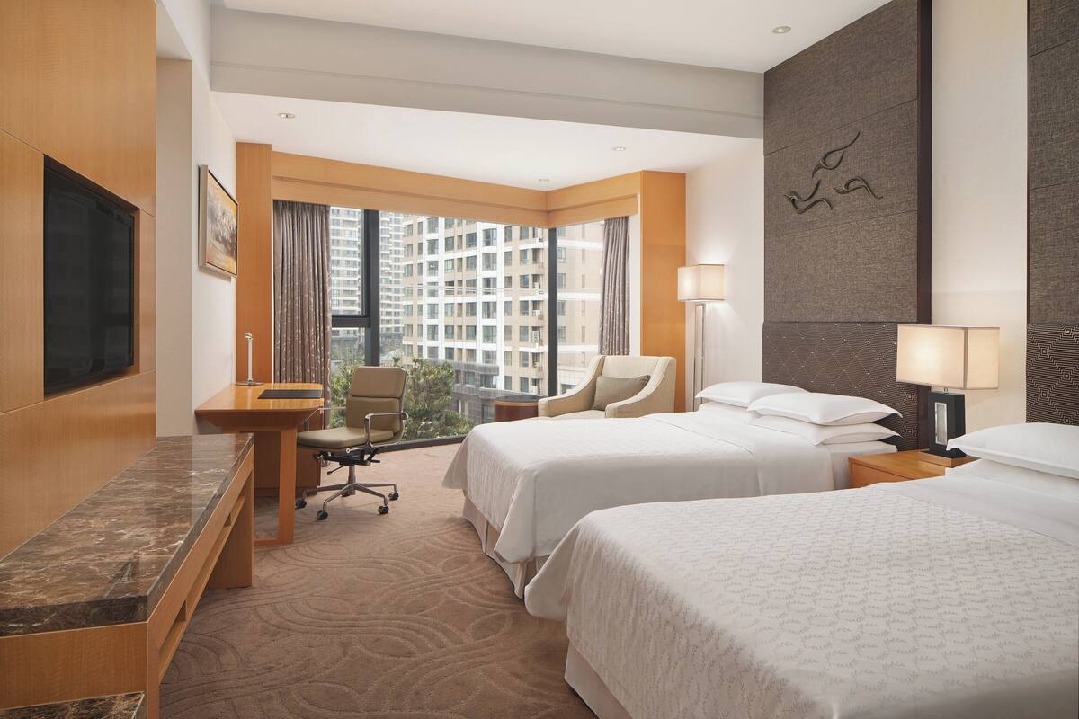 A hotel room with two beds, a desk, a chair, a TV, and a large window with a city view.