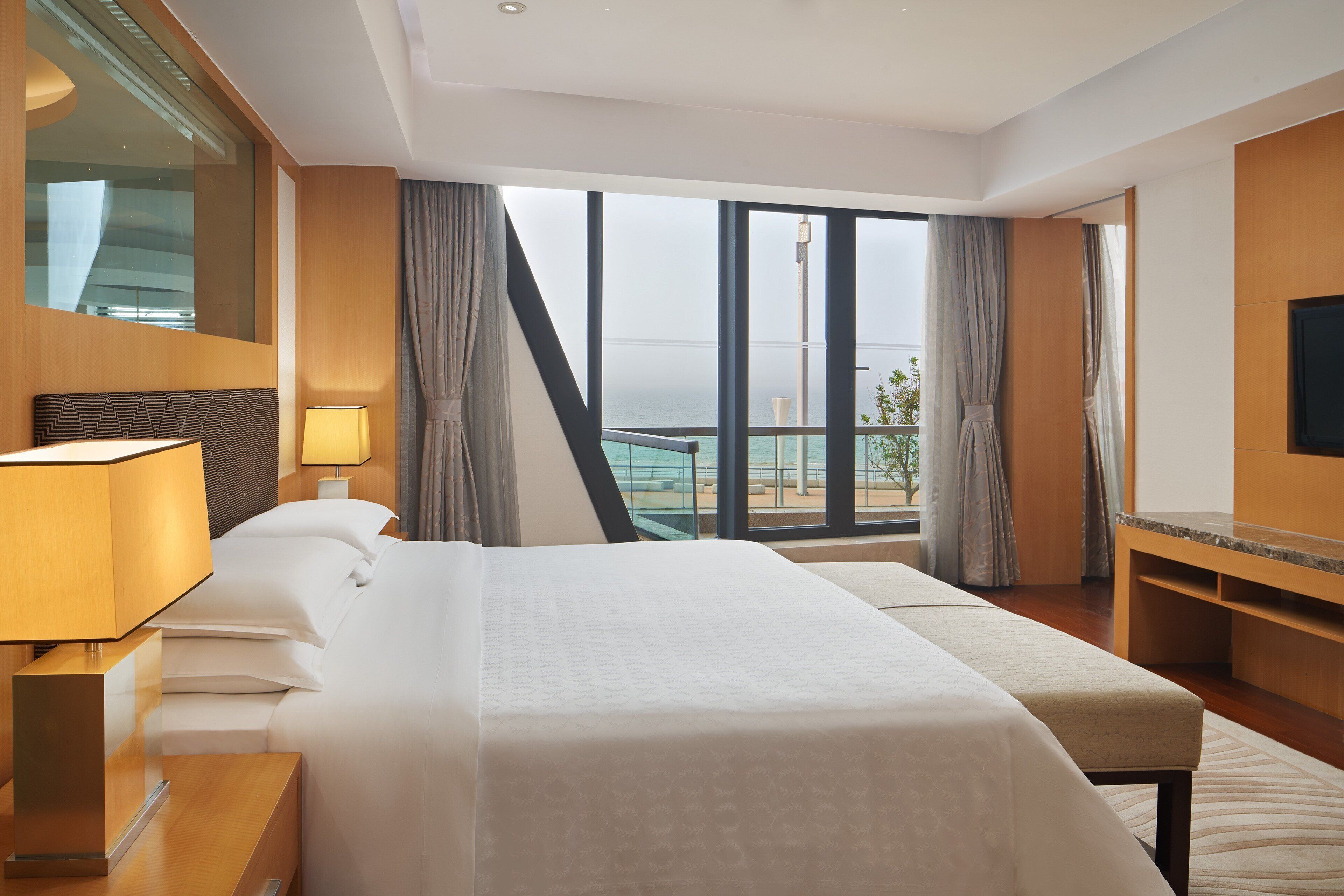 A hotel room with a large bed, a desk, a TV, and a view of the sea.
