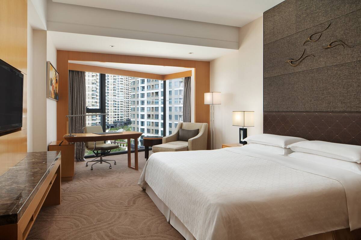 A hotel room with a large bed, a desk, a chair, a TV, and a view of a high-rise building.