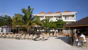Un hotel frente a la playa con múltiples sillones y sombrillas, una zona de piscina y un edificio con techo de tejas y balcones.