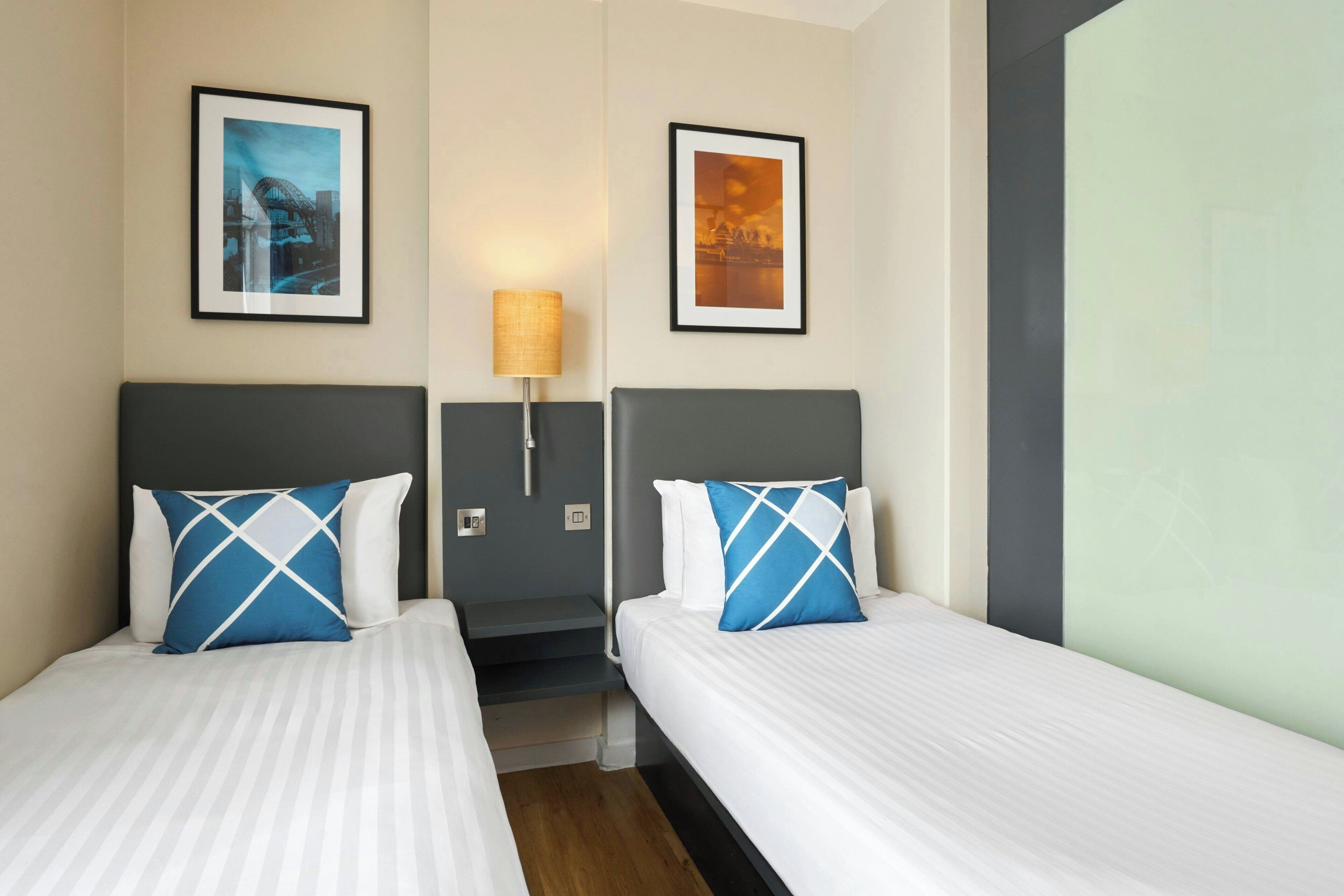 Habitación de hotel con dos camas, una mesita de noche, una lámpara y dos cuadros en las paredes.