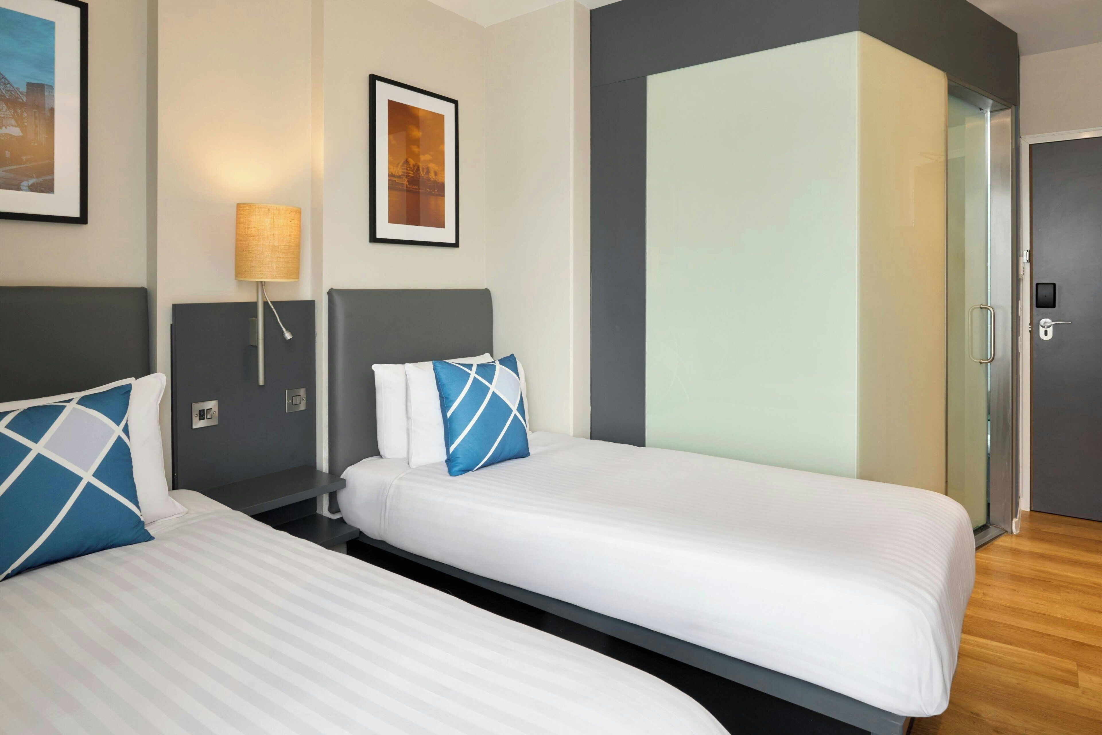 A hotel room with two beds, a bedside table with a lamp, a wardrobe, and framed pictures on the wall.