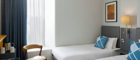 Habitación de hotel con dos camas, un escritorio y una silla, una ventana con cortinas, una lámpara de pared y un cuadro enmarco.