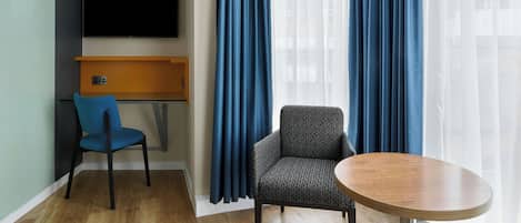 Une chambre d’hôtel équipée d’un téléviseur à écran plat fixé au mur, d’un bureau avec une chaise, d’une table ronde et de deux grandes fenêtres ornées de rideaux bleus.