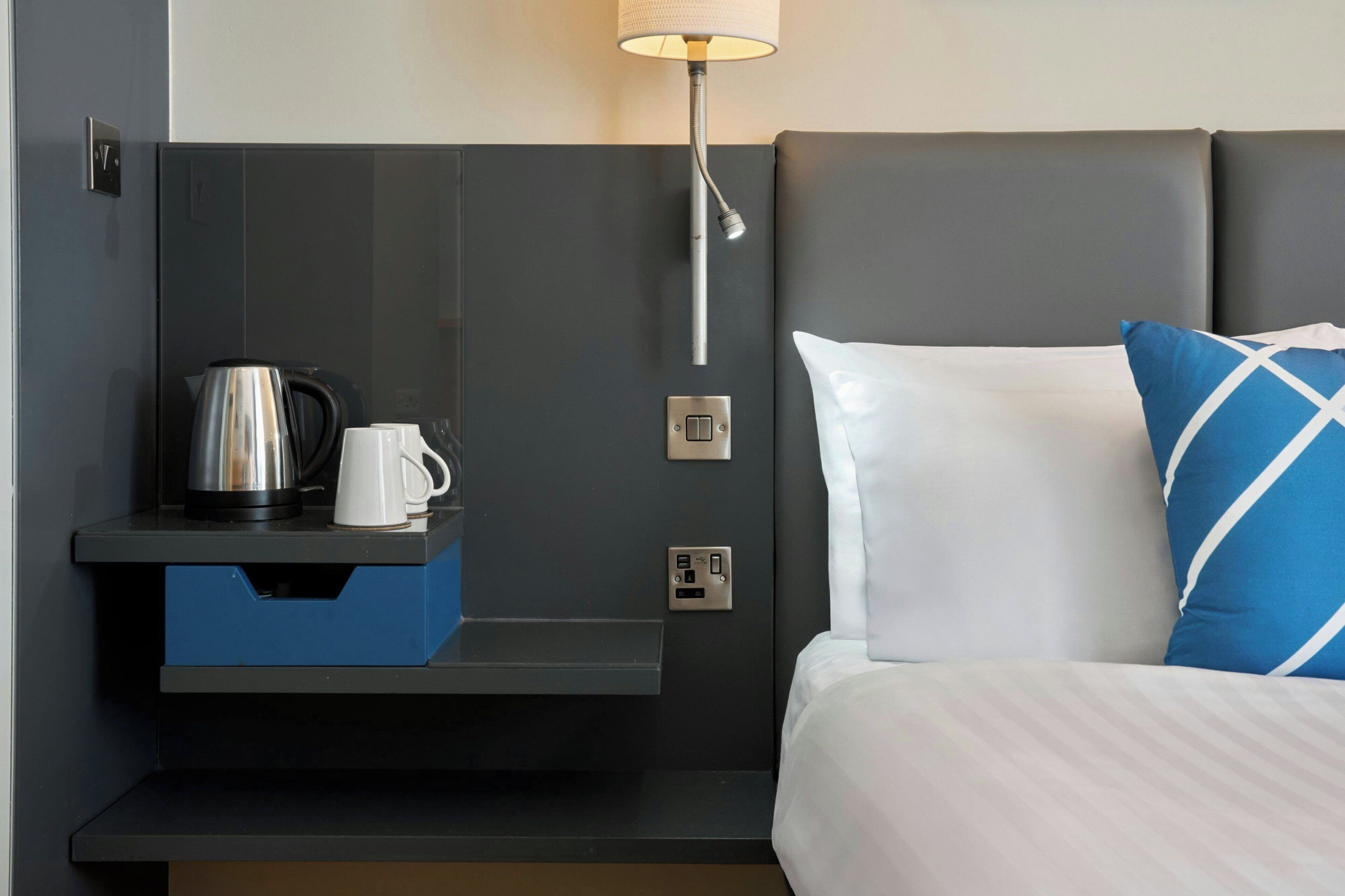 A hotel room with a bed, a nightstand, a lamp, a coffee maker, and a power outlet.