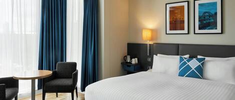 Een hotelkamer met een bed, een stoel, een klein tafeltje, een lamp en twee afbeeldingen in lijsten aan de muur.