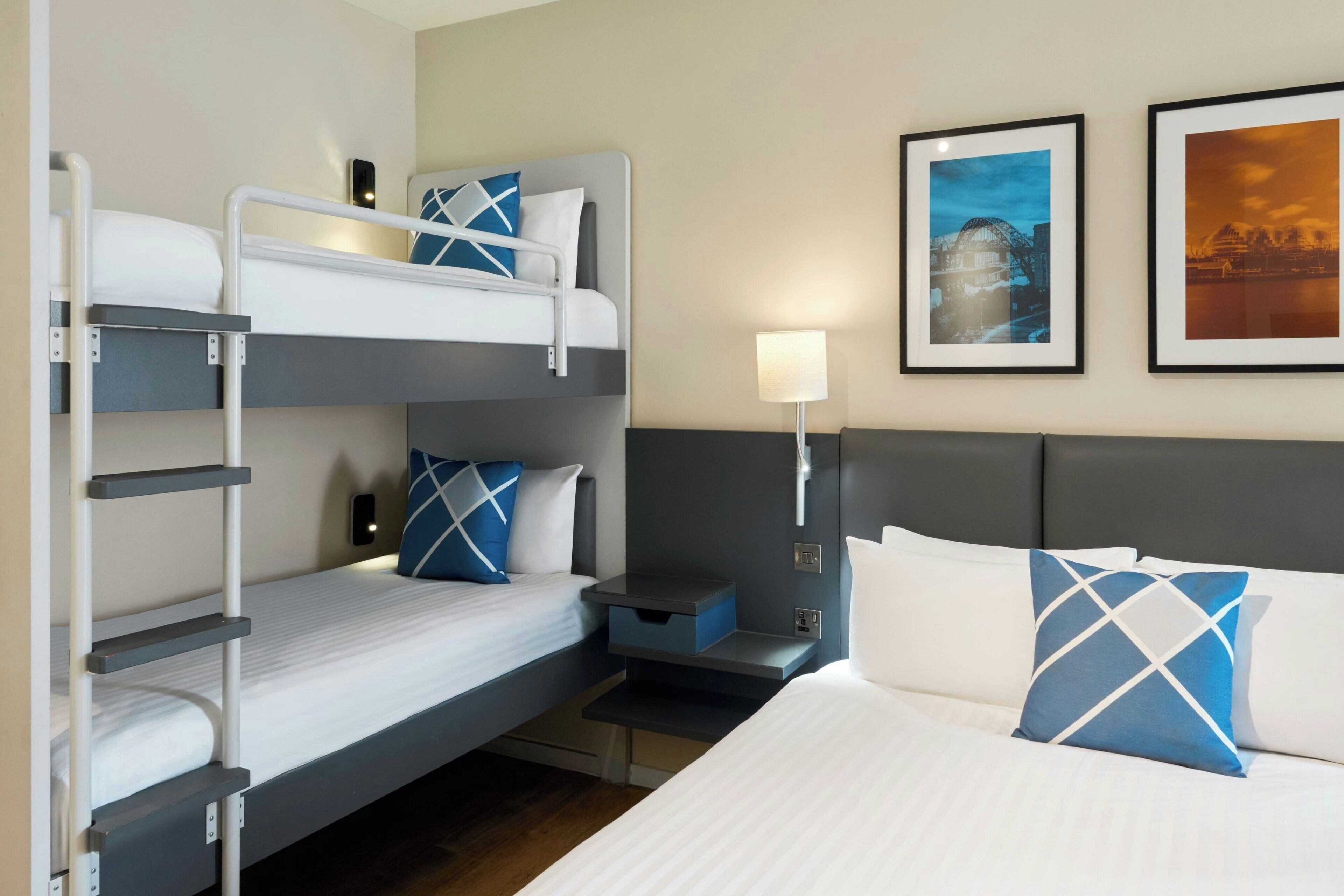 A hotel room with bunk beds, a bedside table, a lamp, and two framed pictures on the wall.