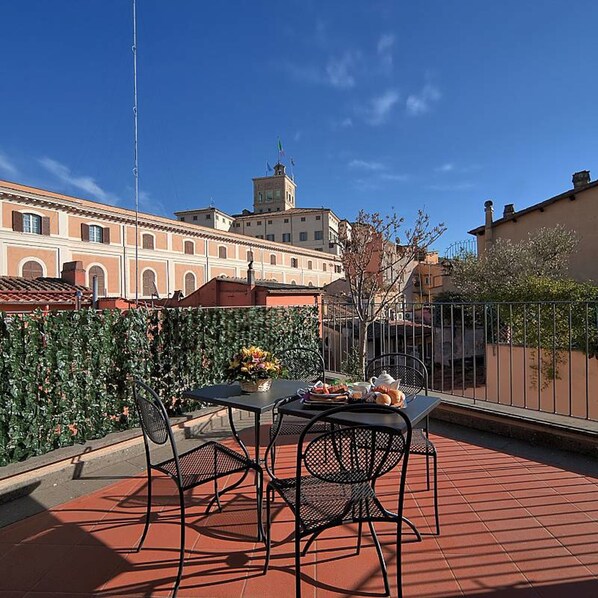 A terrace with a table set for two, surrounded by a metal fence, overlooking a cityscape with historic buildings.