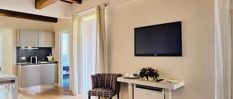 A modern living room with a sofa, a dining area with a table and chairs, a kitchen in the background, and a flat-screen TV mounted on the wall.