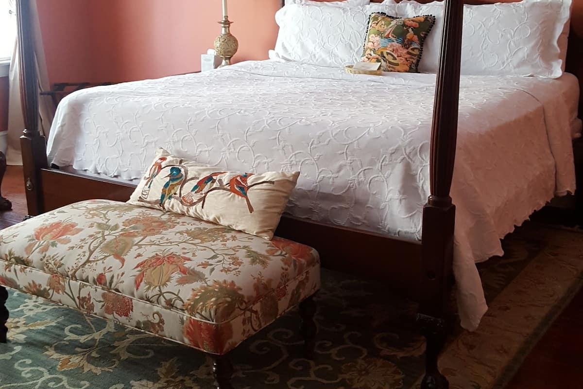 Una camera da letto con un letto a baldacchino, un comodino con una lampada, un pouf floreale e un quadro appeso al muro.