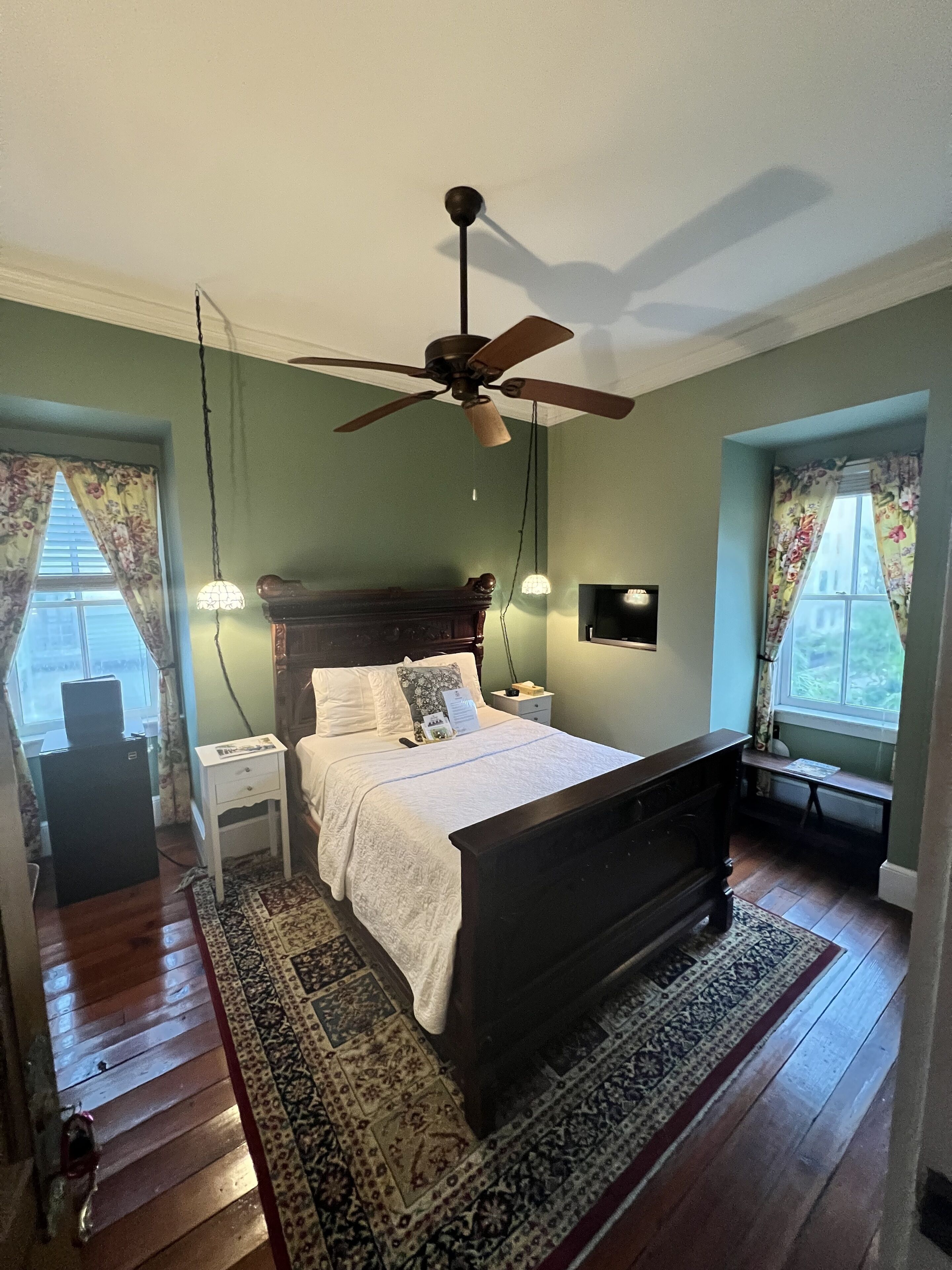 A bedroom with a wooden bed, a ceiling fan, a nightstand, a desk, and a window with curtains.