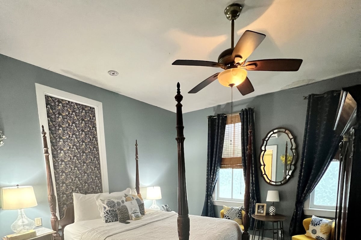 Un letto a baldacchino con ventola a soffitto, un tappeto grande con motivo floreale e vista all'esterno attraverso una finestra con tende.