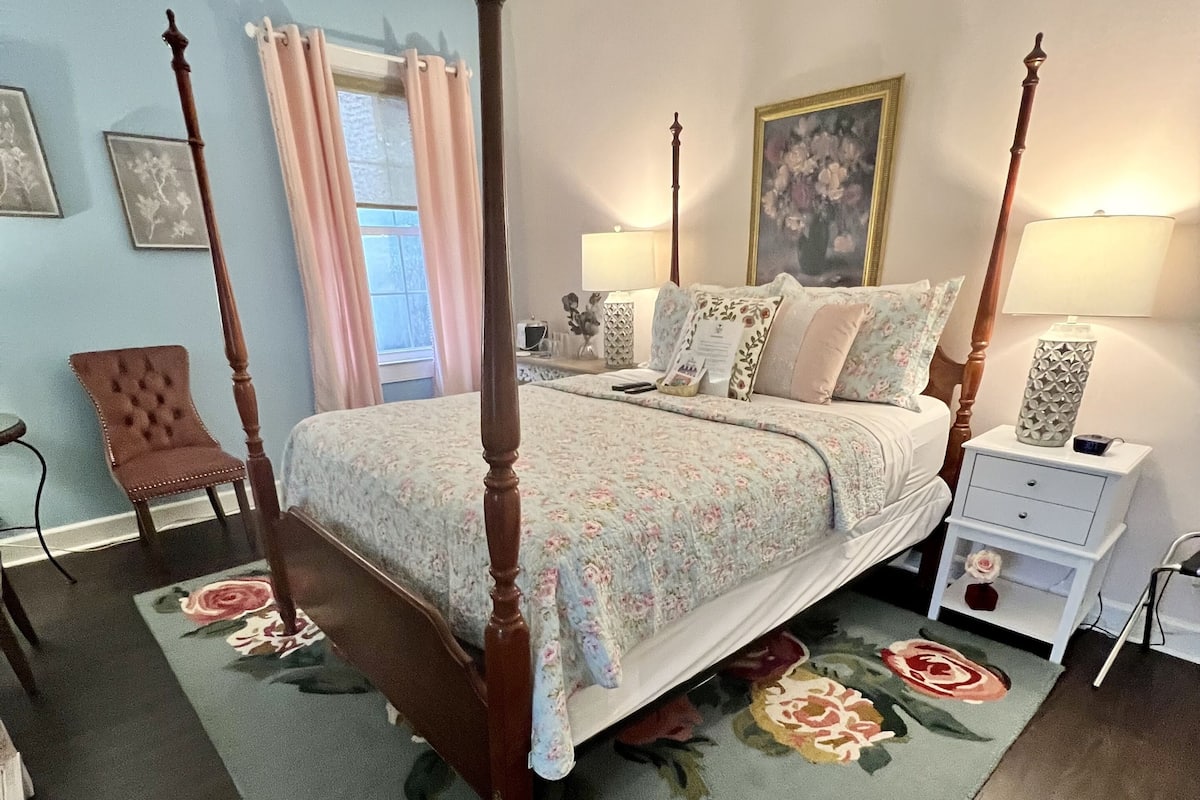 Un letto a baldacchino con biancheria da letto floreale, un comodino con una lampada e una poltrona in una camera da letto con una parete blu e un tappeto floreale.