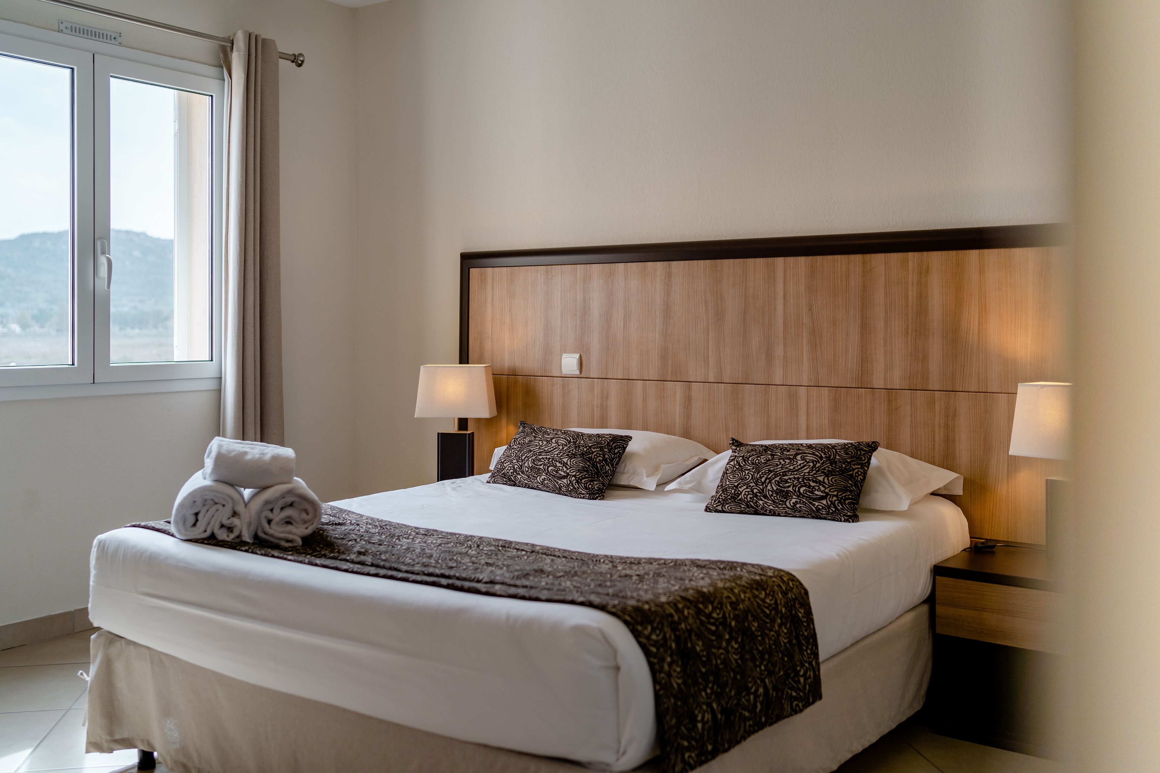 Ein Hotelzimmer mit einem großen Bett, zwei Nachttischlampen, einem hölzernen Kopfteil und einem Fenster mit Vorhängen.