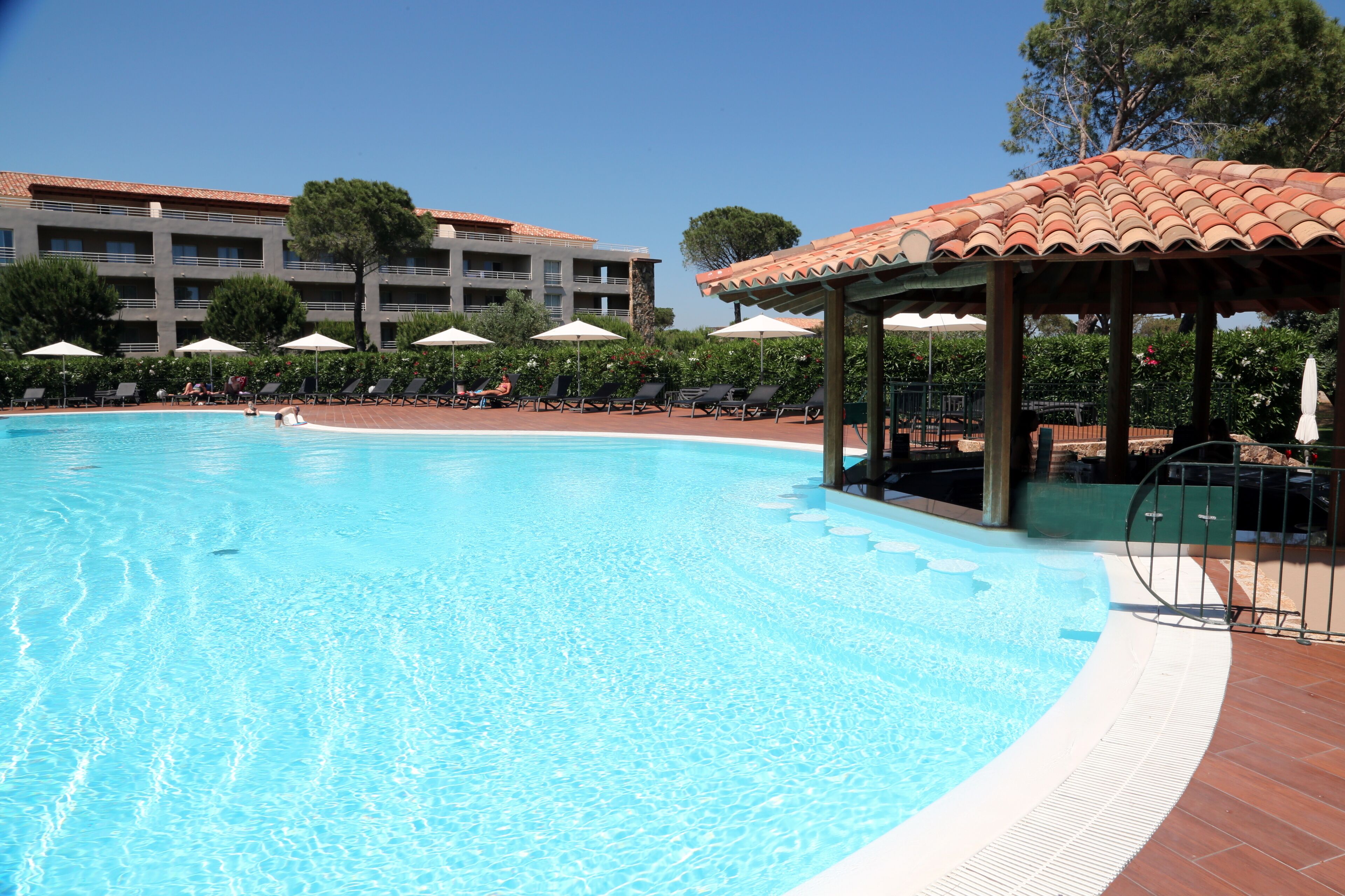 A swimming pool with lounge chairs and umbrellas, a gazebo, and a building with balconies in the background.