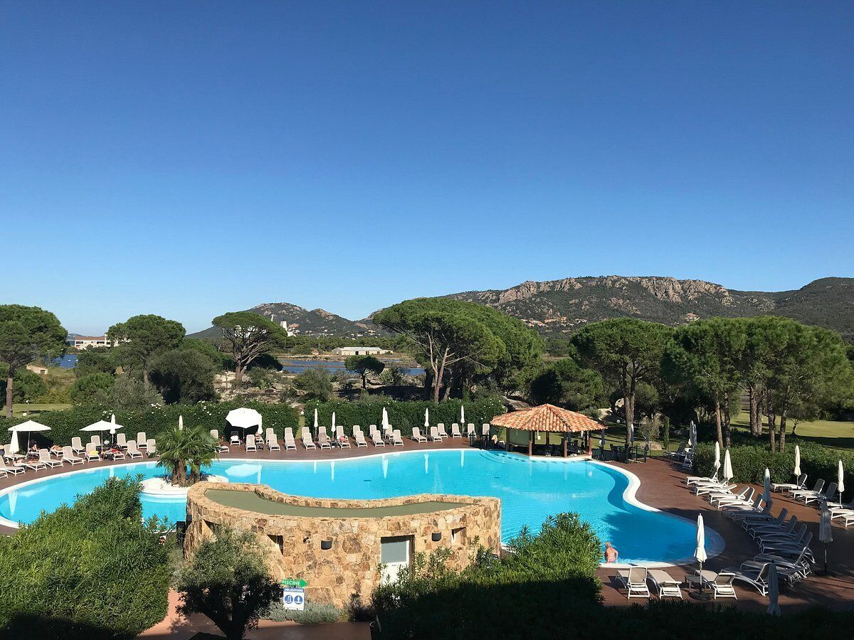 Una piscina grande y curvada con sillas de descanso y sombrillas, rodeada de árboles y montañas al fondo.