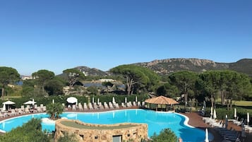 Una piscina grande y curvada con sillas de descanso y sombrillas, rodeada de árboles y montañas al fondo.