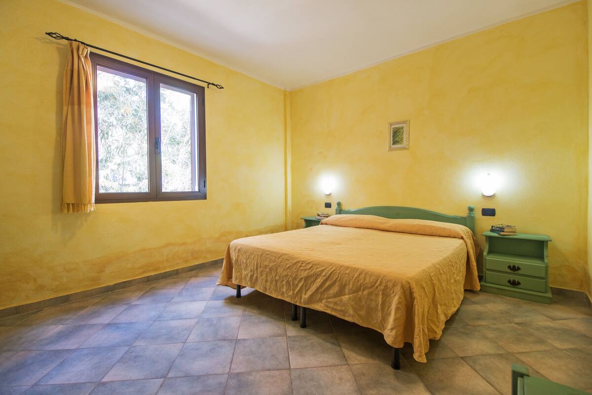Une chambre à coucher comprenant un lit, une table de chevet, une lampe de chevet, une fenêtre avec des rideaux et un luminaire fixé au mur.