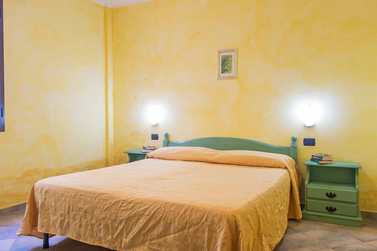 Un lit simple avec une tête de lit et un pied de lit verts, flanqué de deux tables de chevet équipées de tiroirs, dans une pièce aux murs jaunes et avec un seul luminaire suspendu au-dessus du lit.