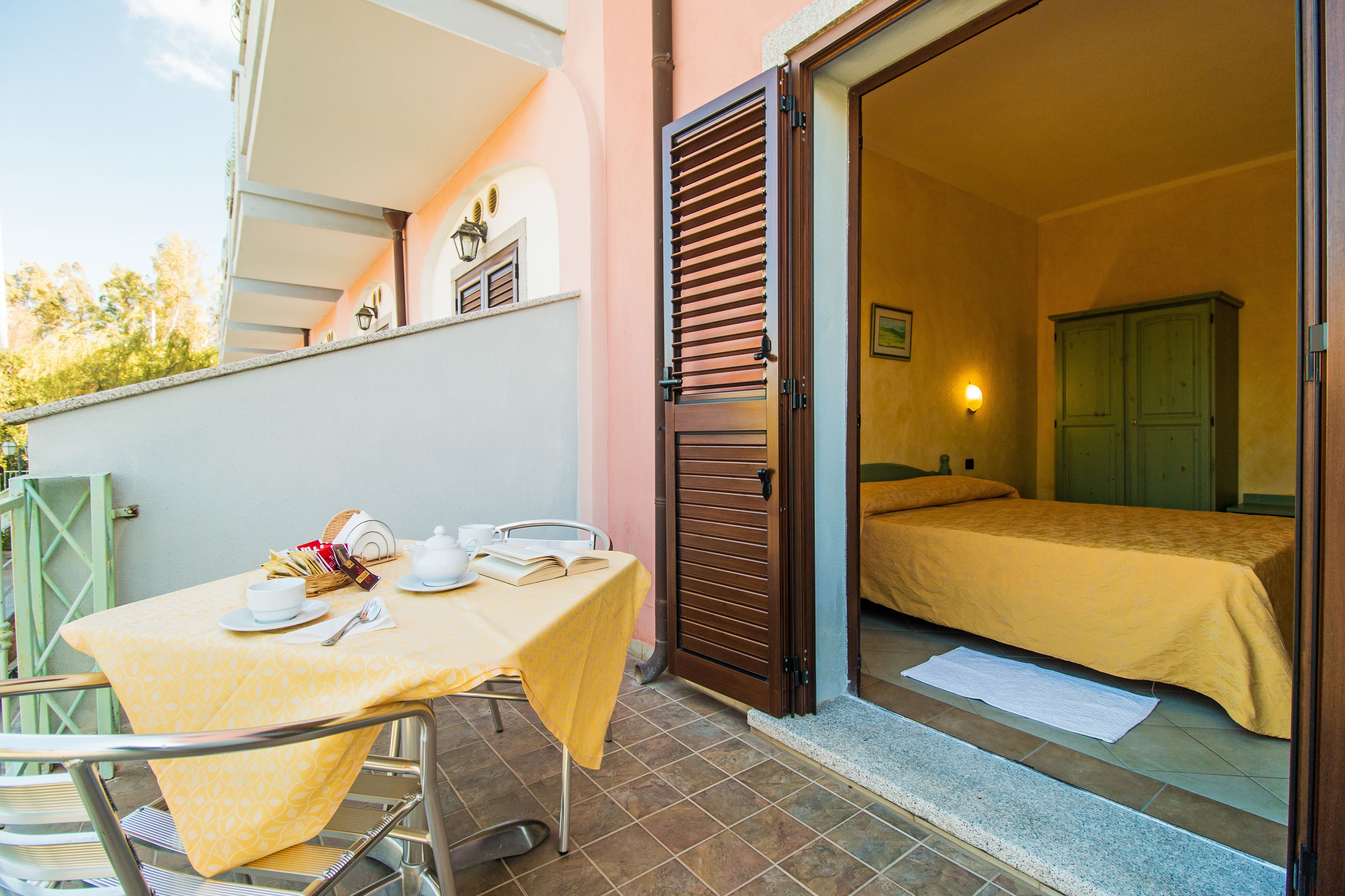 A room with a bed, a table with a yellow tablecloth, and a balcony with a table set for breakfast.