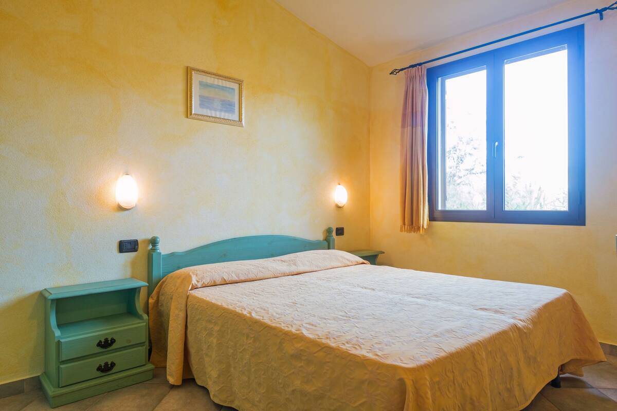 Une chambre à coucher comprenant un lit, deux tables de chevet, une commode, une fenêtre avec des rideaux et un luminaire fixé au mur.