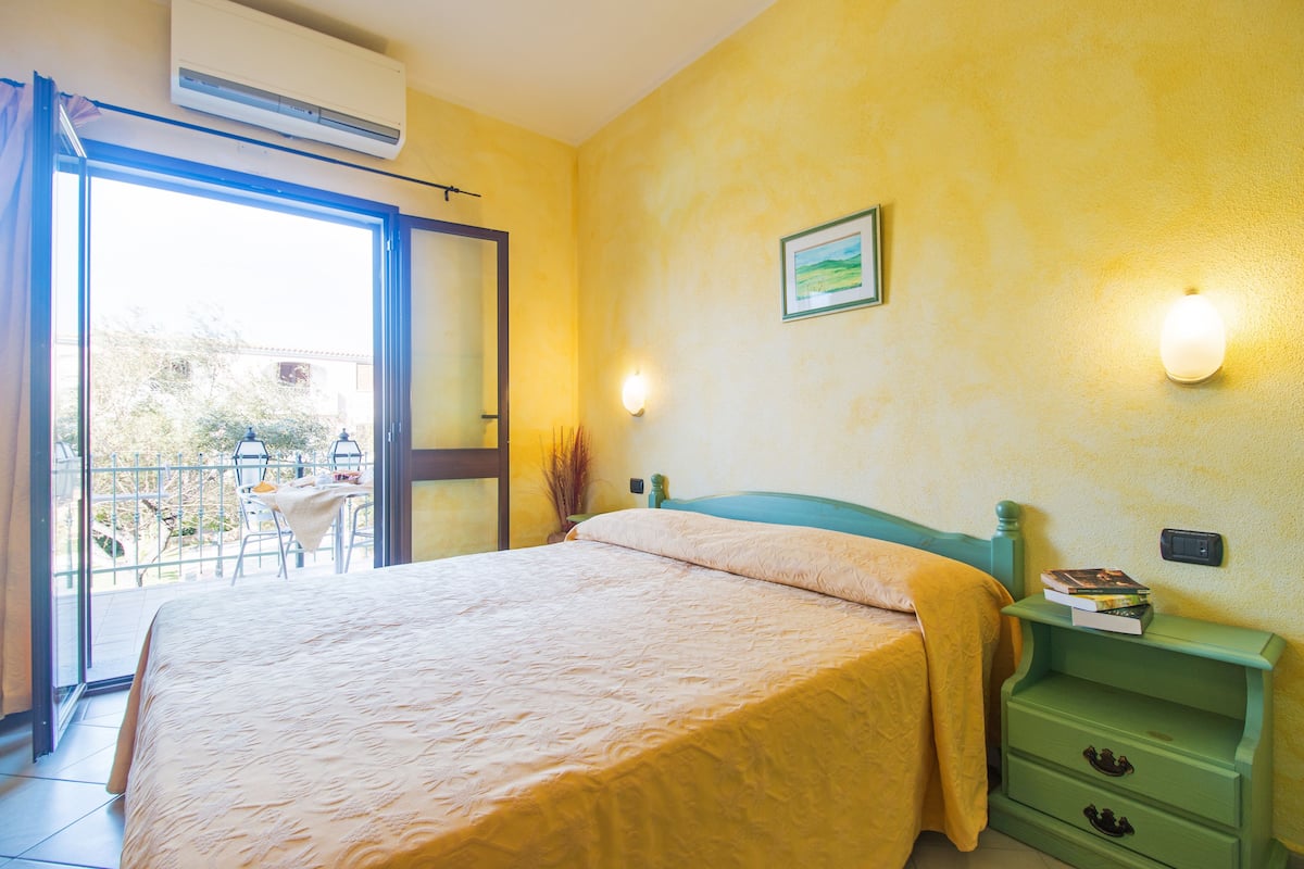Une chambre à coucher avec un lit, une table de chevet verte, une lampe fixée au mur et une fenêtre donnant sur l’extérieur.