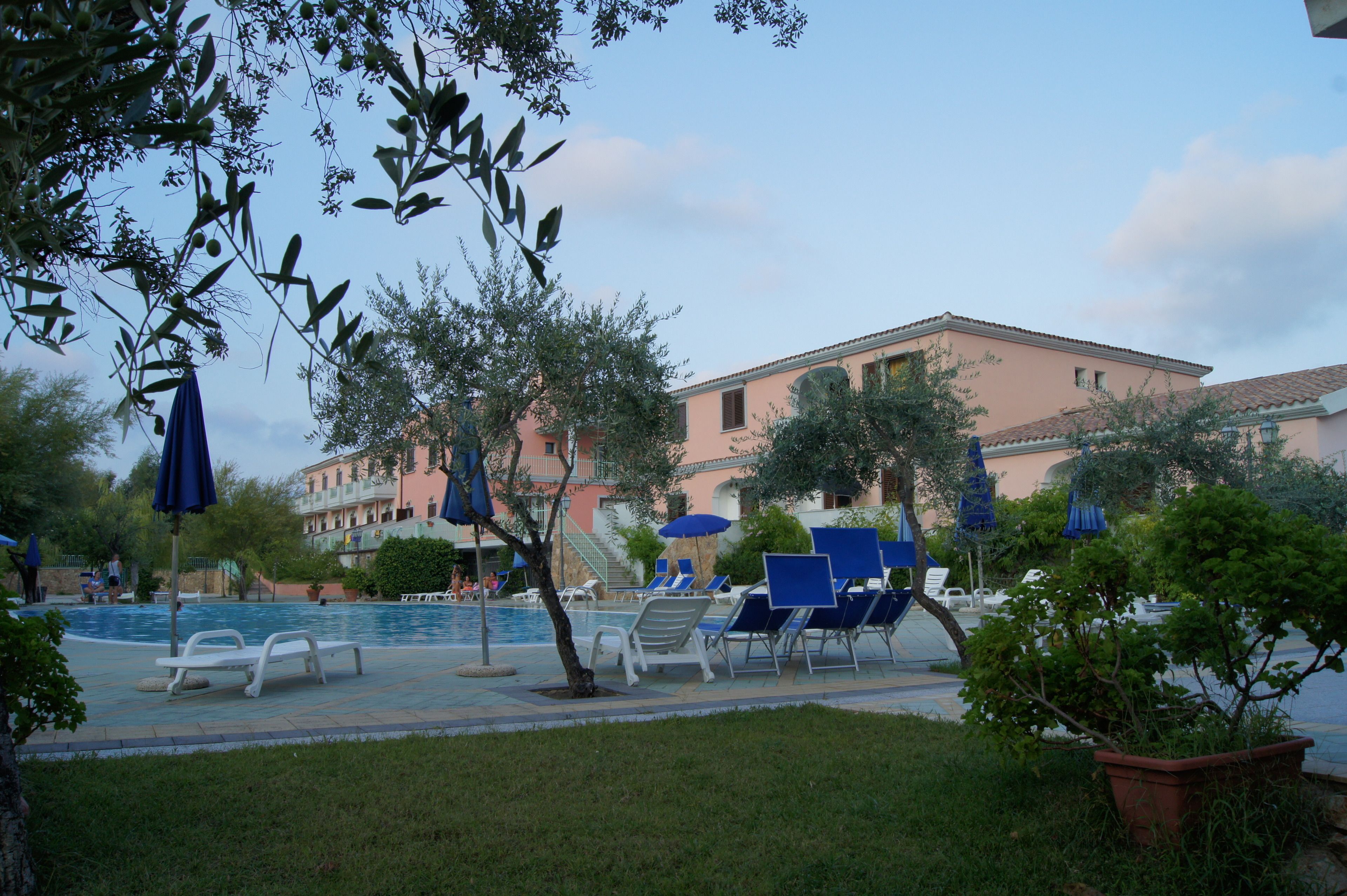 Un espace piscine avec des chaises longues et des parasols, un bâtiment à la façade rose et un ciel dégagé.