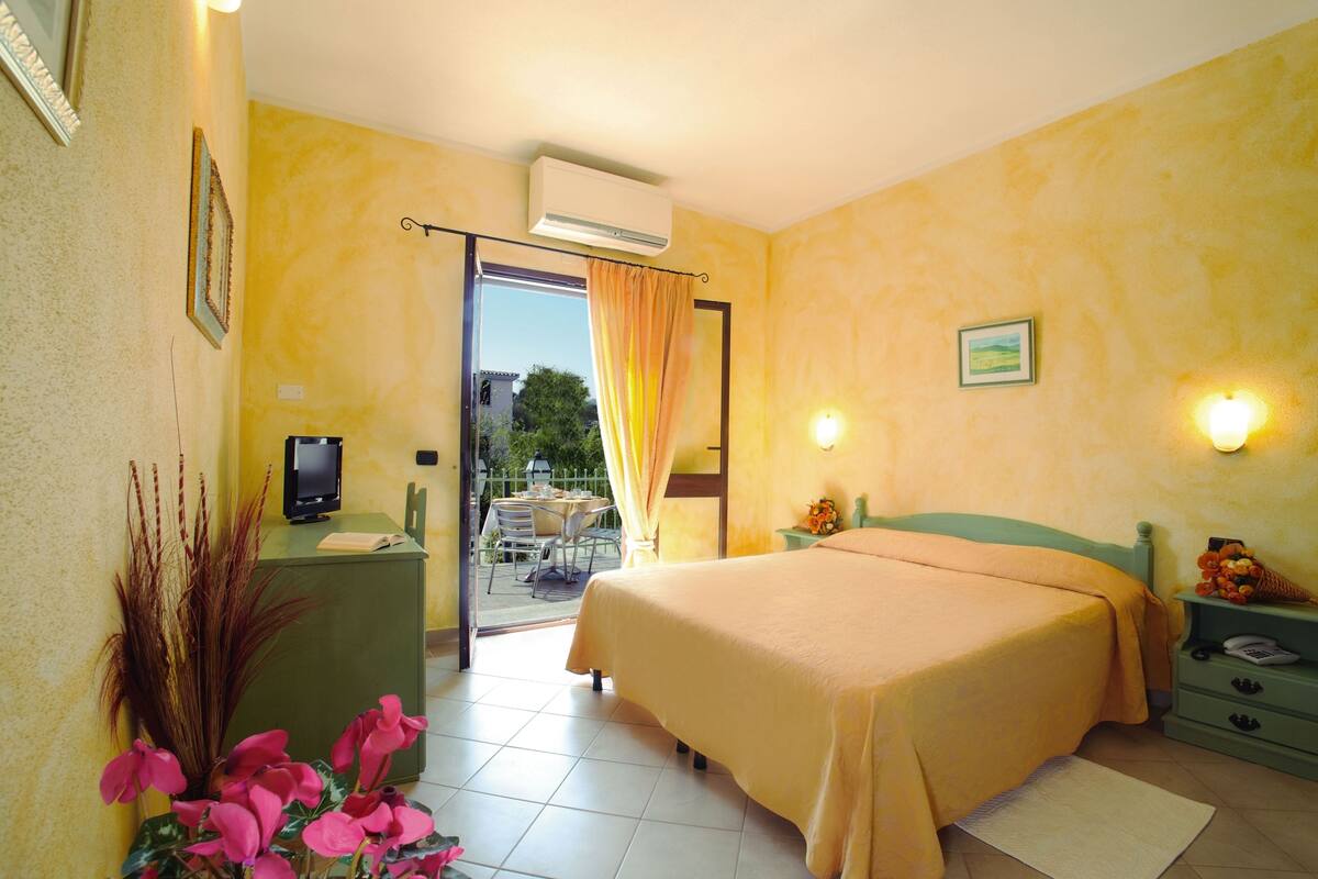 Une chambre d’hôtel comprenant un lit, un bureau avec un ordinateur, une chaise, une petite table avec une chaise, et un balcon avec une table et des chaises.