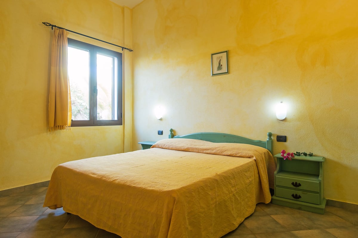 Une chambre à coucher comprenant un lit, une table de chevet, une fenêtre avec des rideaux et un luminaire fixé au mur.