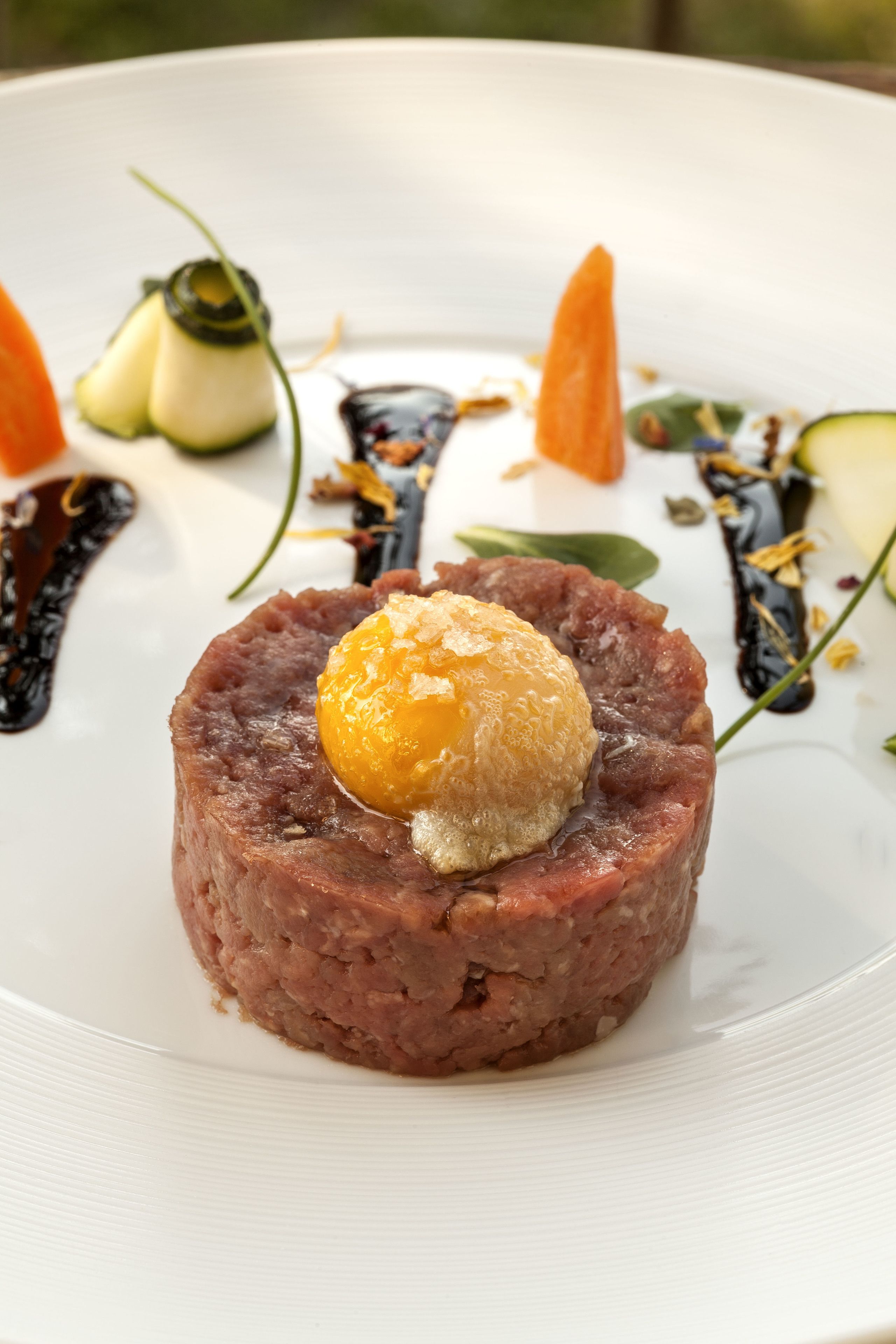 Un plato con una base de carne redonda cubierta por una esfera amarilla, rodeada de guarniciones sobre un plato blanco.