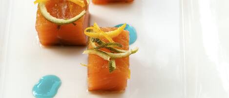 Un plato rectangular blanco con cinco porciones de tartar de salmón adornado con ralladura de limón y hierbas, y puntos de salsa azul.