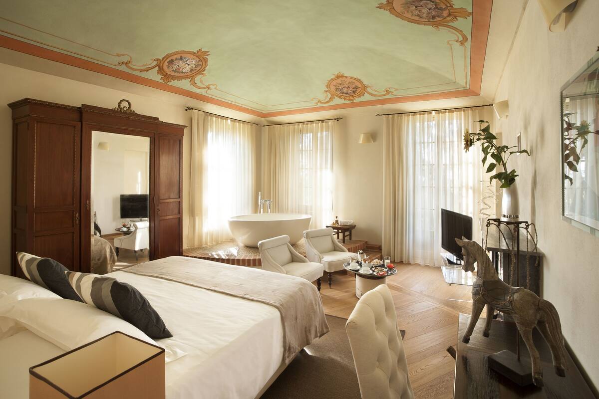 Habitación de hotel con una cama grande, dos sillones, una mesita, una bañera y un televisor.