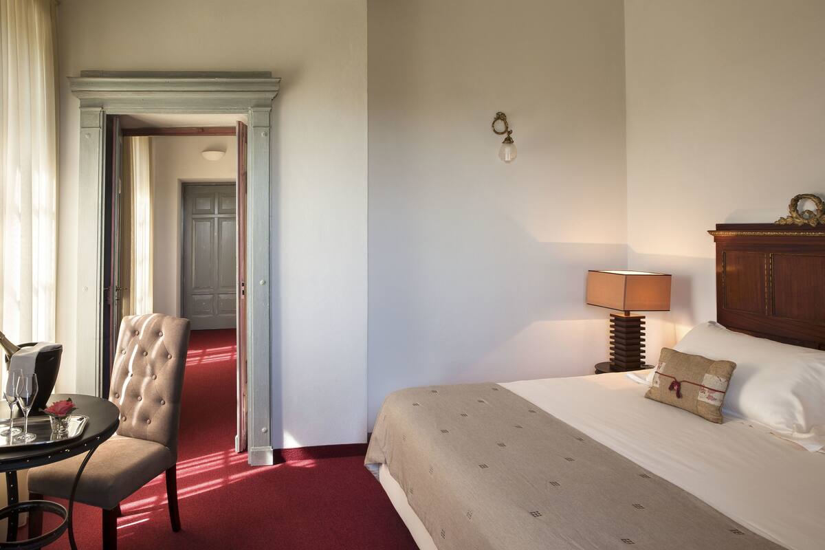 Una habitación de hotel con una cama, una silla, una mesa con una bandeja y vasos, y una lámpara.