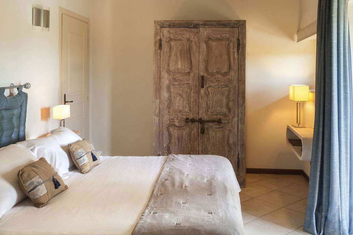 Un dormitorio con cama, armario de madera, mesita de noche con lámpara y ventana con cortinas.