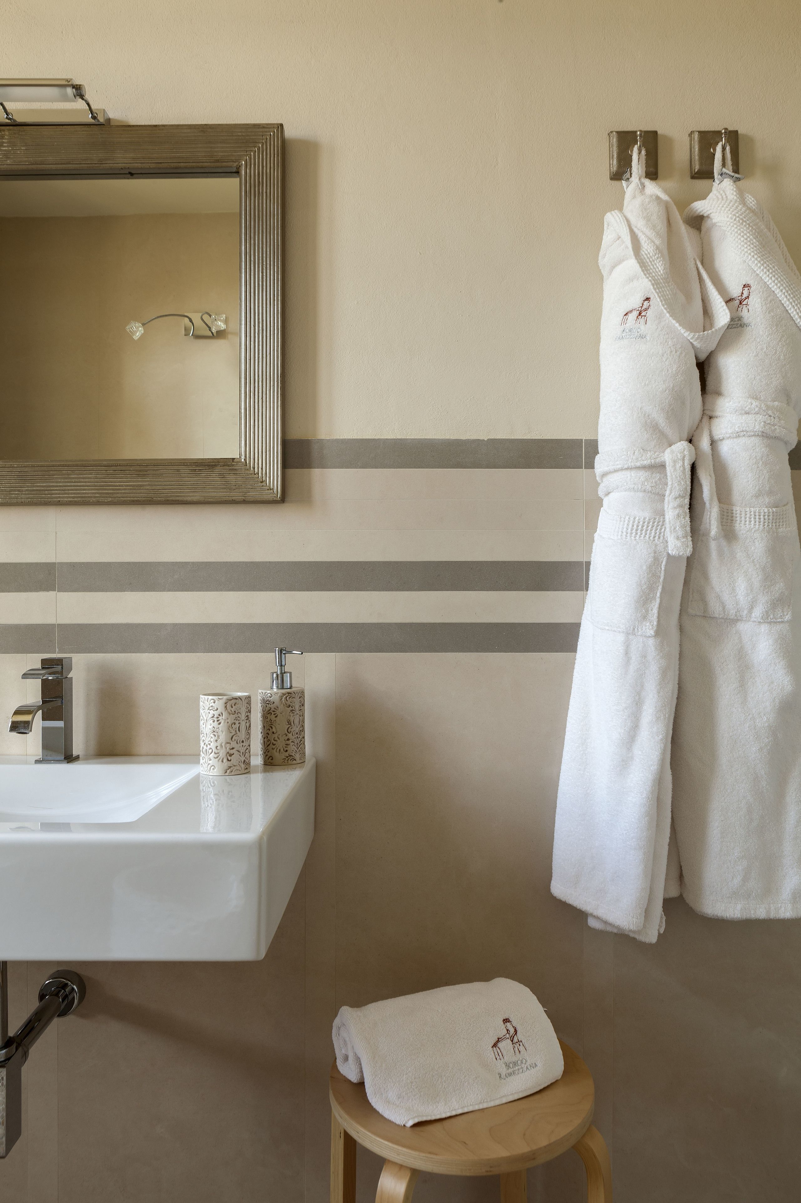 Un bagno con un lavandino bianco, uno specchio e due asciugamani bianchi appesi a ganci.