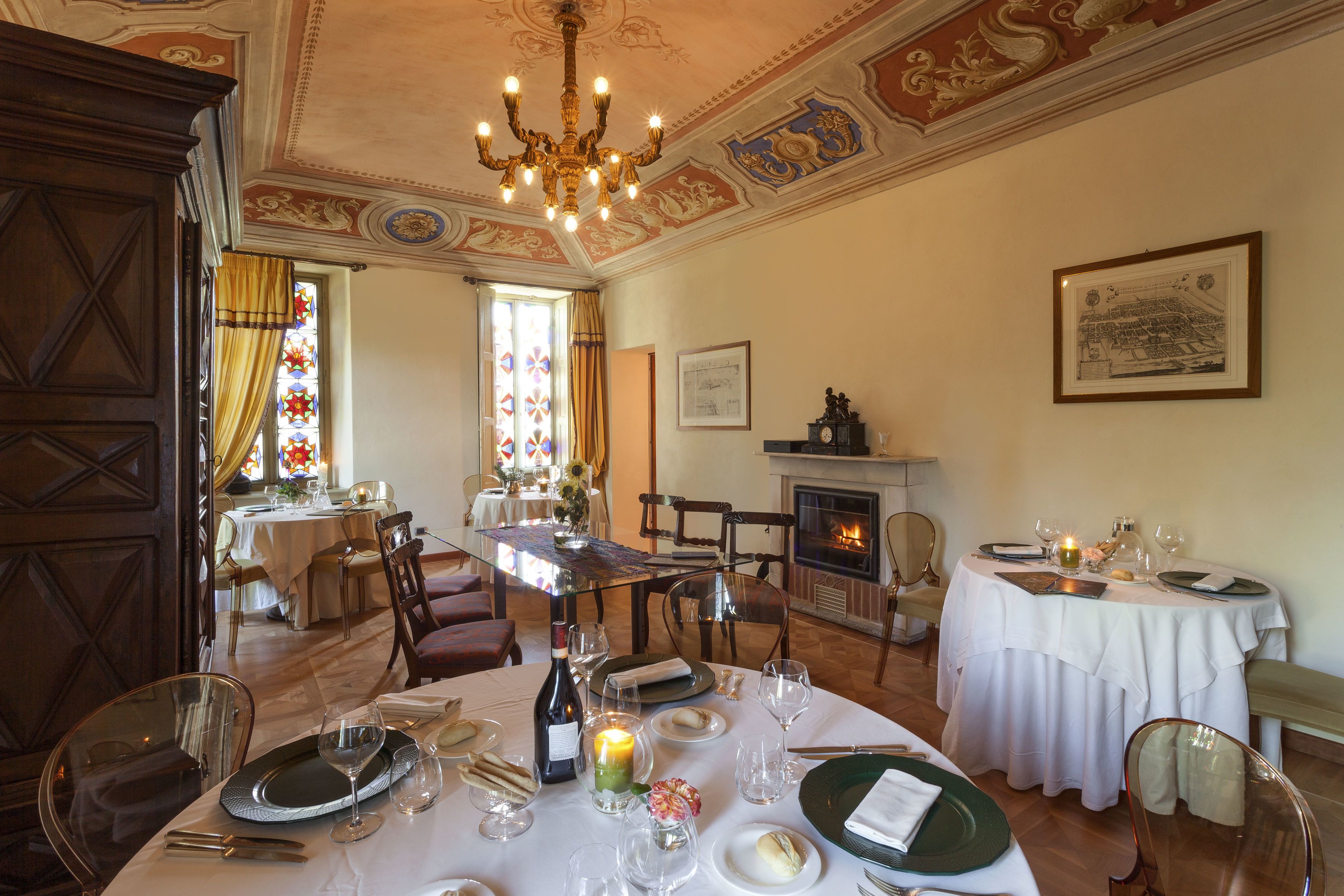 Uma área de refeições com mesas redondas, cadeiras e um candelabro. Há uma lareira e uma janela de vitrais ao fundo.